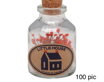 Peut inclure: Un bocal en verre rempli de 100 punaises rouges et blanches. Le bocal a un couvercle en liège et une étiquette qui dit "Little House" avec une petite illustration de maison et le texte "Produced by KINKAME".