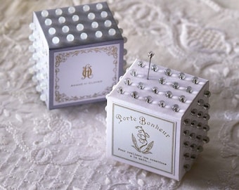 Peut inclure: Deux coussinets à épingles blancs et gris avec un design décoratif. Les coussinets à épingles sont recouverts de petites perles blanches et portent une étiquette qui indique "Porte Bonheur" et "Pour réaliser vos créations à la main..."