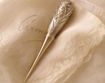 Peut inclure: Un dé à coudre en argent avec un motif de muguet. Le dé à coudre est posé sur un tissu blanc avec une bordure en dentelle.