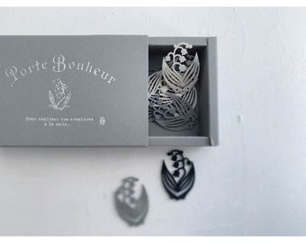 Peut inclure: Une petite boîte grise avec le texte "Porte Bonheur" et la phrase "Pour réaliser vos créations à la main..." imprimée sur le dessus. La boîte est ouverte et contient plusieurs découpes en papier blanc de fleurs de muguet. Il y a aussi deux découpes de muguet sur la table, une blanche et une noire.