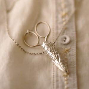 May include: A silver necklace with a pair of scissors charm and a lily of the valley pendant. The necklace is on a white fabric background.