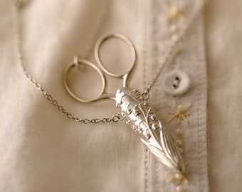 Peut inclure: Un collier en argent avec un pendentif en forme de ciseaux et un pendentif en forme de muguet. Le collier est sur un fond en tissu blanc.