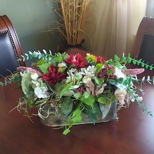 Könnte beinhalten: Rechteckiges Blumenarrangement mit roten, weißen und grünen Blumen in einem verwitterten Behälter. Das Arrangement steht auf einem dunkelbraunen Tisch. Der Hintergrund umfasst zwei Stühle und eine Vase mit hohen, getrockneten Pflanzen.