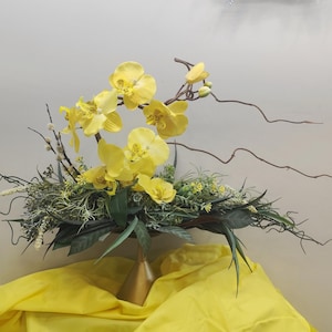 Può includere: Composizione floreale con vibranti orchidee gialle, accentuata da fogliame verde e delicati rami marroni. L'arrangiamento è posto su uno sfondo di tessuto giallo, creando un contrasto sorprendente. I fiori sono il punto focale.