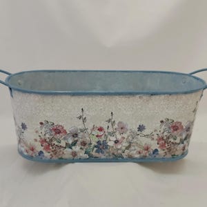 Könnte beinhalten: Ovale Metallpflanzschale mit hellblauem Rand und Griffen. Die Pflanzschale hat ein Blumendesign mit rosa, lila und blauen Blumen auf einem strukturierten weißen Hintergrund. Ein dekorativer Behälter für Pflanzen.