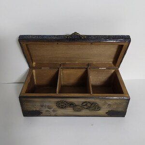May include: Open wooden box with three compartments, featuring a steampunk design. The box has a dark stained finish with decorative metal gears and corner accents. The lid is open, revealing the interior.