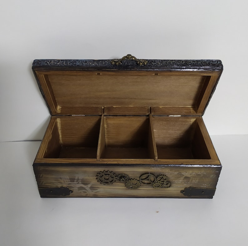 May include: Open wooden box with three compartments, featuring a steampunk design. The box has a dark stained finish with decorative metal gears and corner accents. The lid is open, revealing the interior.