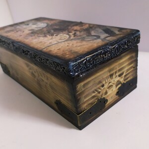 May include: A rectangular wooden box with a hinged lid. The box has a distressed, aged appearance with a dark brown finish. The lid features a vintage-style design, and the edges are trimmed with ornate black metal. The corners are reinforced with metal.