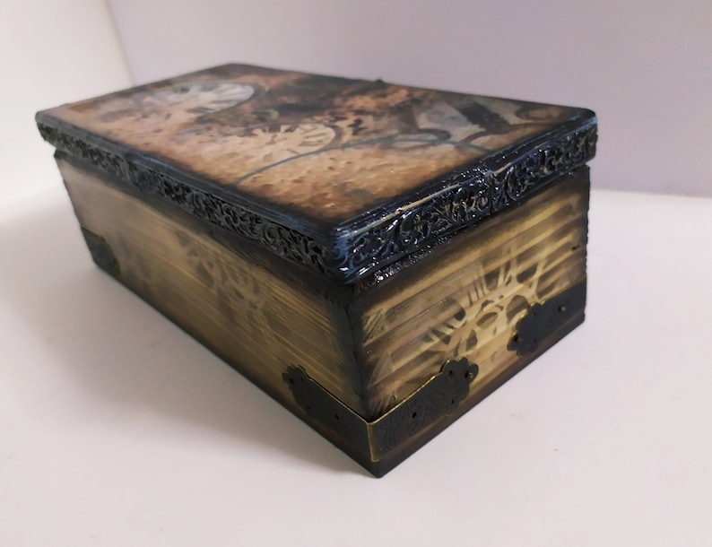 May include: A rectangular wooden box with a hinged lid. The box has a distressed, aged appearance with a dark brown finish. The lid features a vintage-style design, and the edges are trimmed with ornate black metal. The corners are reinforced with metal.
