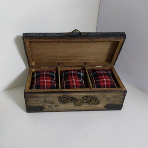 May include: Open wooden box with three compartments, each holding a red and black plaid pillow. The box has a dark stained finish, metal corner accents, and decorative gears. The lid is open, revealing the interior.