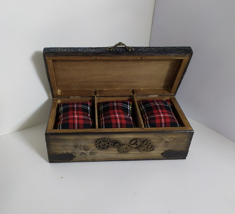 May include: Open wooden box with three compartments, each holding a red and black plaid pillow. The box has a dark stained finish, metal corner accents, and decorative gears. The lid is open, revealing the interior.