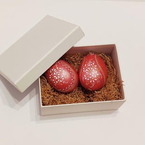 Könnte beinhalten: Zwei rote dekorative Ostereier mit weißen Blumenmustern in einer weißen Schachtel. Die Eier liegen in braunem, strohartigem Material, wobei der Deckel der Schachtel leicht geöffnet ist. Die Eier sind etwa 7.6 cm hoch.
