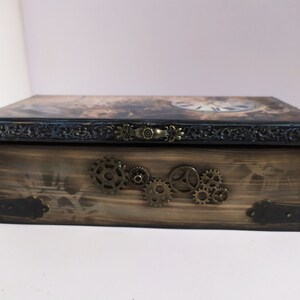 May include: Wooden box with a steampunk design. The box features a clock face on the lid, decorative metal gears, and ornate metal hardware. The box is stained in shades of brown and black.