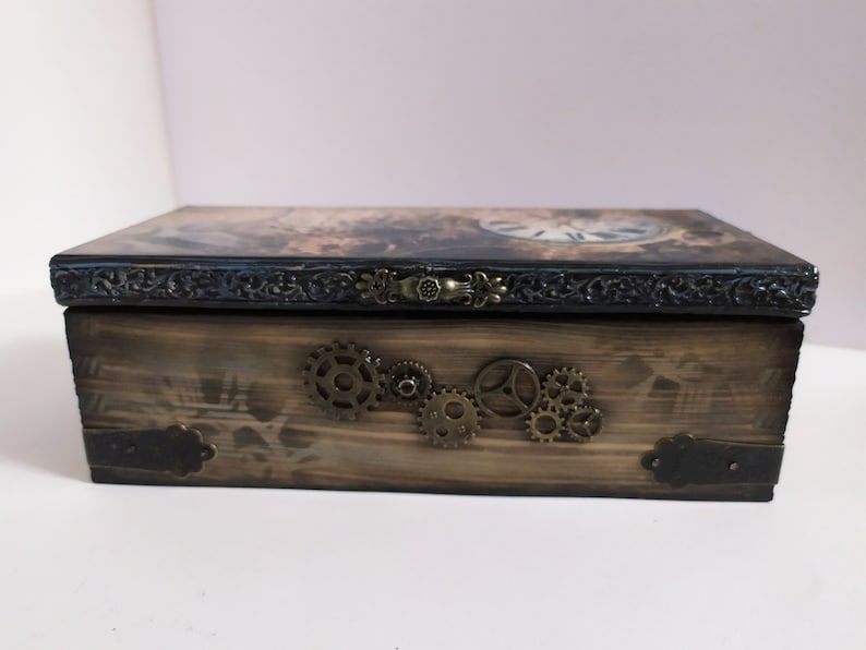 May include: Wooden box with a steampunk design. The box features a clock face on the lid, decorative metal gears, and ornate metal hardware. The box is stained in shades of brown and black.