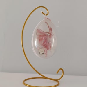Könnte beinhalten: Ein dekoratives, eiförmiges Ornament mit einer rosa Ballerina in einem transparenten Gehäuse, das an einem goldfarbenen Metallständer hängt. Am Fuß der Ballerina befindet sich eine weiße Rose. Der Ständer steht auf einer weißen Oberfläche.