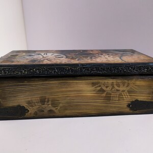 May include: Wooden box with a clock face design on the lid. The box has a dark, ornate border and metal corner accents. The wood has a distressed, aged appearance, with a decorative pattern on the front.