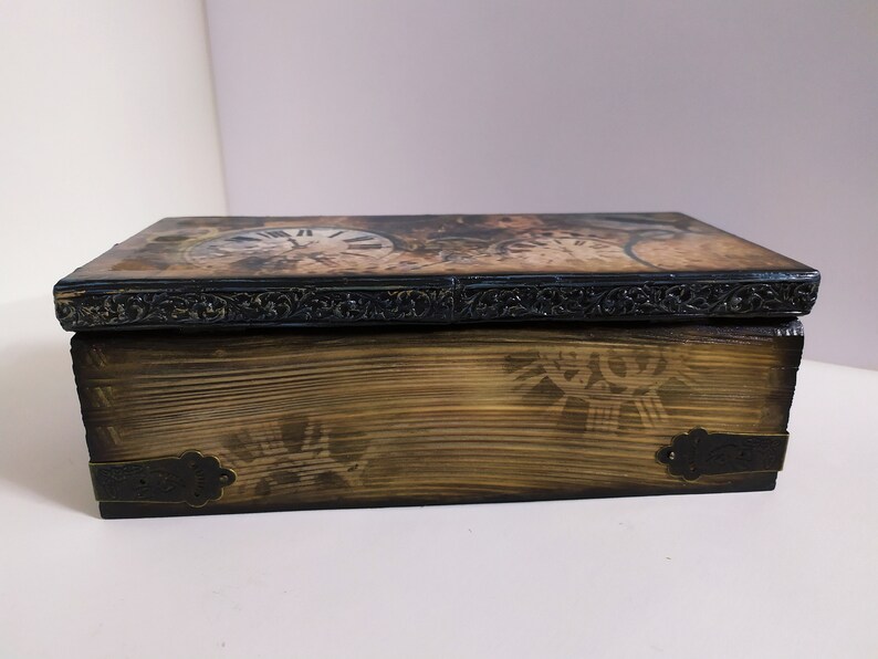 May include: Wooden box with a clock face design on the lid. The box has a dark, ornate border and metal corner accents. The wood has a distressed, aged appearance, with a decorative pattern on the front.