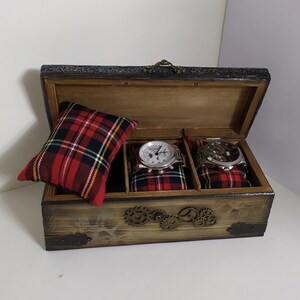 May include: Wooden watch box with a hinged lid, featuring a distressed finish and decorative metal accents. The interior holds two watches on plaid cushions and a separate plaid pillow. The box has a steampunk aesthetic.