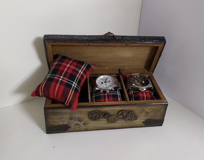 May include: Wooden watch box with a hinged lid, featuring a distressed finish and decorative metal accents. The interior holds two watches on plaid cushions and a separate plaid pillow. The box has a steampunk aesthetic.