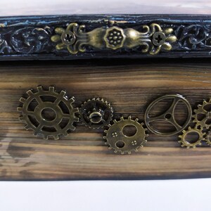 May include: Wooden box with a steampunk aesthetic. The box features a dark stained wood finish, ornate metal hardware, and decorative brass gears. The top edge has a dark blue and black trim with decorative carvings.