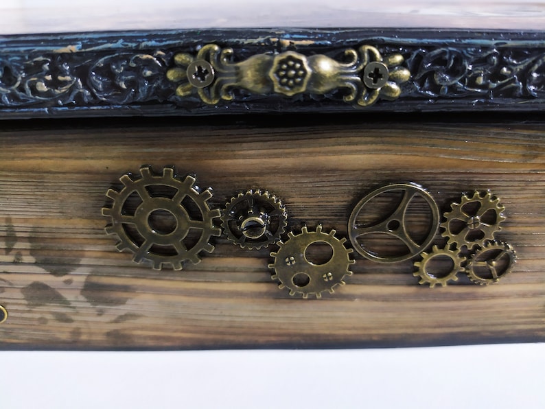 May include: Wooden box with a steampunk aesthetic. The box features a dark stained wood finish, ornate metal hardware, and decorative brass gears. The top edge has a dark blue and black trim with decorative carvings.