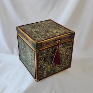 May include: A square decorative box with an intricate pattern in green, gold, and red. The box has a tassel and gold trim. It is likely used for storage or display.