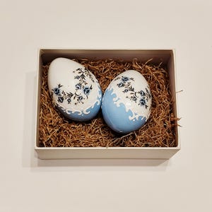 Può includere: Due uova di Pasqua decorative in una scatola bianca riempita di paglia. Le uova sono bianche con disegni floreali blu e una base azzurra. Le uova sono di forma ovale e hanno una finitura lucida. La scatola è rettangolare e dal design semplice.