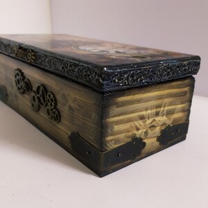 May include: A rectangular wooden box with a steampunk design. The box is stained brown with black accents and metal gear embellishments. The lid features a decorative border and a detailed image. The box is approximately 10 inches long.