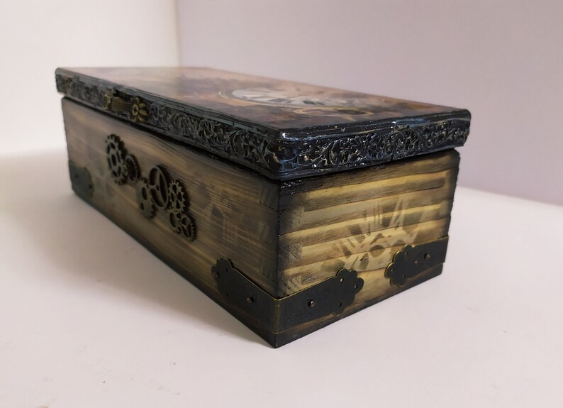 May include: A rectangular wooden box with a steampunk design. The box is stained brown with black accents and metal gear embellishments. The lid features a decorative border and a detailed image. The box is approximately 10 inches long.
