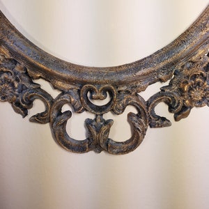 May include: A close-up of the bottom of an oval-shaped ornate picture frame. The frame is made of a dark, distressed metal with gold accents. The frame has a decorative, scroll-like design.