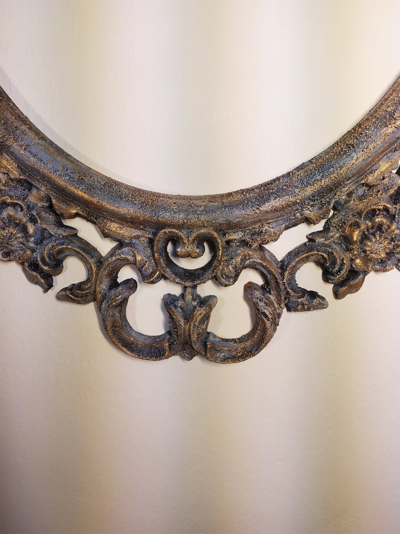 May include: A close-up of the bottom of an oval-shaped ornate picture frame. The frame is made of a dark, distressed metal with gold accents. The frame has a decorative, scroll-like design.