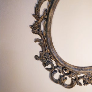 May include: A close-up of an ornate, oval-shaped picture frame with a dark, aged finish. The frame has intricate, floral-inspired details and a slightly distressed look.