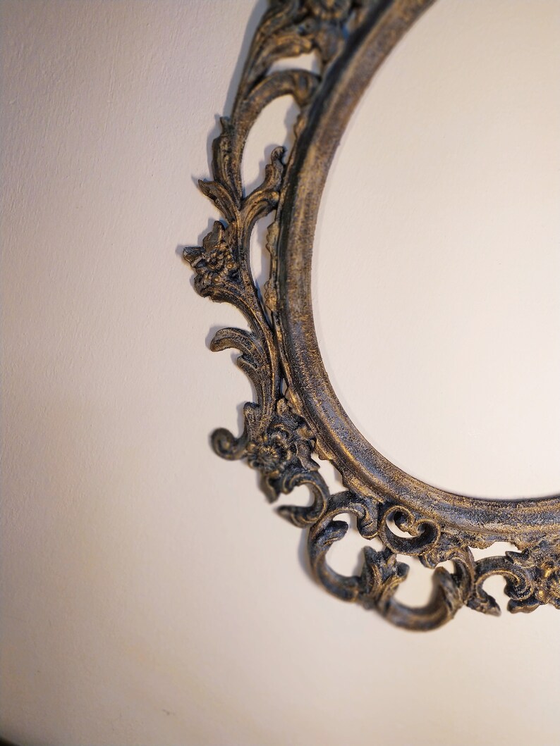 May include: A close-up of an ornate, oval-shaped picture frame with a dark, aged finish. The frame has intricate, floral-inspired details and a slightly distressed look.