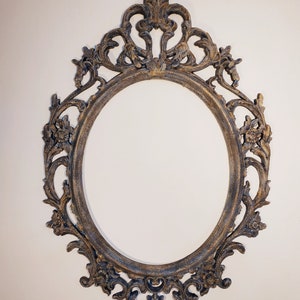 May include: An ornate oval-shaped picture frame with a distressed gold finish. The frame has intricate details and a decorative top.