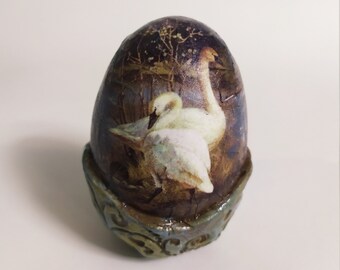 Puede incluir: Un objeto decorativo en forma de huevo con una imagen pintada de dos cisnes blancos sobre un fondo oscuro. El huevo descansa sobre una base decorativa con detalles azules y dorados. Tiene un aspecto vintage o antiguo.