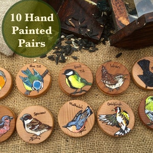May include: Ten hand-painted wooden bird ornaments, each featuring a different species of bird, including a robin, blue tit, great tit, chaffinch, sparrow, nuthatch, wren, blackbird, goldfinch, and greenfinch. The ornaments are arranged in two rows on a hessian background.