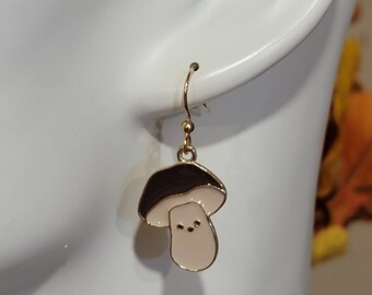 May include: Gold mushroom-shaped dangle earrings with a black top and a light pink bottom. The mushroom has a small, white face with two black dots for eyes.