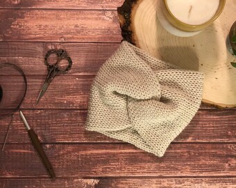 May include: A light beige knitted headband with a twisted knot design, resting on a wooden surface. A pair of decorative scissors, a crochet hook, and a candle are also visible. The headband is a cozy accessory.