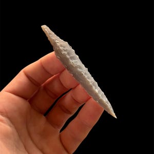 Op de afbeelding: Een grijze, met de hand gekapte pijlspits met een scherpe punt en een getextureerd oppervlak. Het oude artefact wordt tegen een zwarte achtergrond gehouden, wat het gedetailleerde vakmanschap en de historische betekenis laat zien. De pijlspits is lichtgrijs.