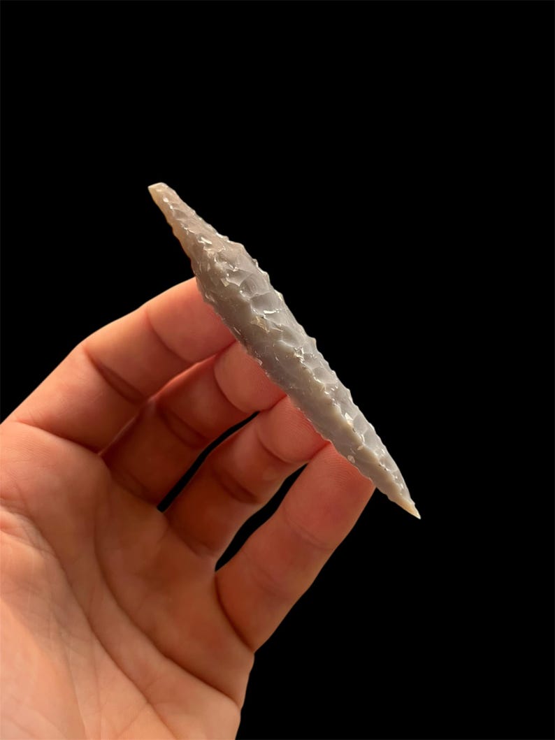 Op de afbeelding: Een grijze, met de hand gekapte pijlspits met een scherpe punt en een getextureerd oppervlak. Het oude artefact wordt tegen een zwarte achtergrond gehouden, wat het gedetailleerde vakmanschap en de historische betekenis laat zien. De pijlspits is lichtgrijs.