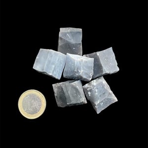 Puede incluir: Un conjunto de seis cristales en bruto de color azul claro, de forma irregular. Una moneda de euro se encuentra junto a los cristales para comparar el tamaño. Fondo negro.