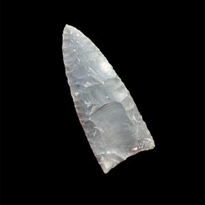 Puede incluir: Una punta de flecha de sílex gris translúcido, con una punta afilada y una superficie rugosa. De forma triangular, parece haber sido tallada a mano, mostrando la artesanía antigua. El fondo es negro.