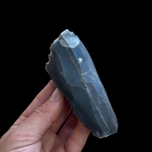 Op de afbeelding: Een ruwe, donkergrijze vuursteen met een glad, conchoïdaal breukvlak. Het mineraalexemplaar vertoont subtiele kleurnuances en een scherpe, grillige rand aan één kant, wat de natuurlijke, ongepolijste en onregelmatige geologische vorm benadrukt.