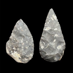 May include: Two gray flint hand axes with a teardrop shape are displayed against a black background. The axes have a rough, textured surface with lighter-colored inclusions. They are likely prehistoric tools.