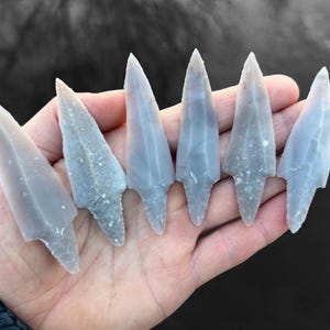 以下が含まれることがあります： 鋭い先端と質感のある表面を持つ、6つの灰色のフリント製の矢じり。 矢じりは一列に並べられ、その職人技を示しています。 背景はぼやけた濃い灰色です。