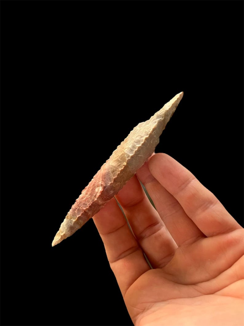 Op de afbeelding: Een handgehouden, bleke stenen pijl- of speerpunt. De stenen tool heeft een scherpe, puntige punt en een textuur met roodbruine en beige tinten. Het artefact wordt tegen een zwarte achtergrond gehouden.