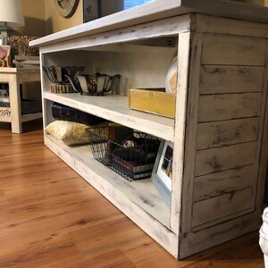 May include: White distressed wood media console with three shelves. The console has a distressed white finish and is decorated with various items, including a pillow, a basket, and a picture frame.