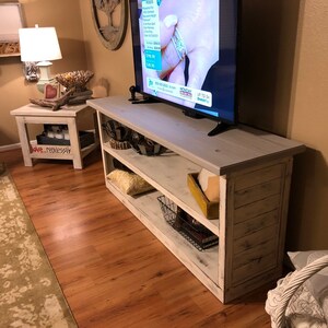 May include: Whitewashed wood TV stand with two shelves, a flat screen TV on top, and a basket and books on the bottom shelf. The stand has a distressed white finish and is in a living room setting.