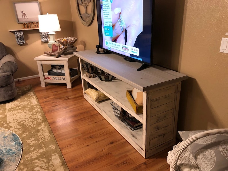 May include: Whitewashed wood TV stand with two shelves, a flat screen TV on top, and a basket and books on the bottom shelf. The stand has a distressed white finish and is in a living room setting.