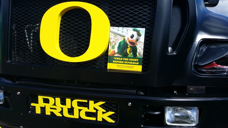 May include: A black truck with a yellow "O" on the front grill. The truck has a yellow "DUCK TRUCK" on the front bumper. A book titled "Twas the Night Before Duckmas" is attached to the grill.